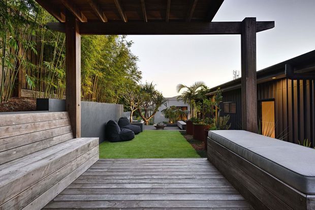 A contemporary outdoor courtyard and deck framed by a dark timber pergola. The space includes built-in weathered timber bench seating, a large cushioned built-in daybed, artificial grass, black lounge seating, raised planters, bamboo screening and tropical planting, with dark-clad structures surrounding the courtyard.