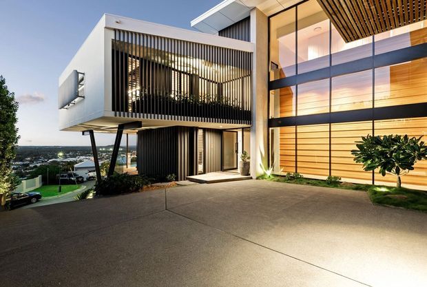 A contemporary multi-level residence viewed from the driveway at dusk. The home features crisp white rendered volumes, extensive floor-to-ceiling glazing, dark vertical screening and framing, warm timber wall lining, a cantilevered upper floor, and a broad concrete driveway. Landscape planting and a distant suburban outlook are visible around the entry.