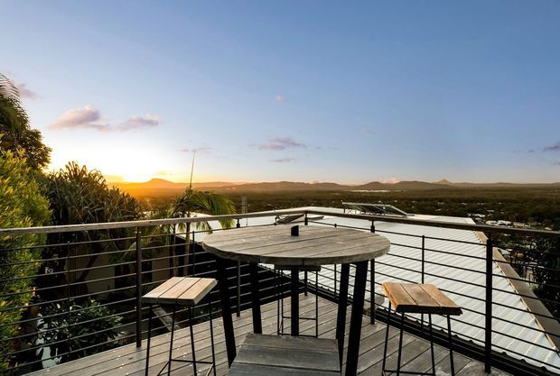 Elevated outdoor terrace or balcony with a dark timber deck, minimalist black metal cable railing, and a round outdoor bar table with stools. The terrace overlooks a broad green landscape and distant hills at sunset, creating an expansive, relaxed setting.