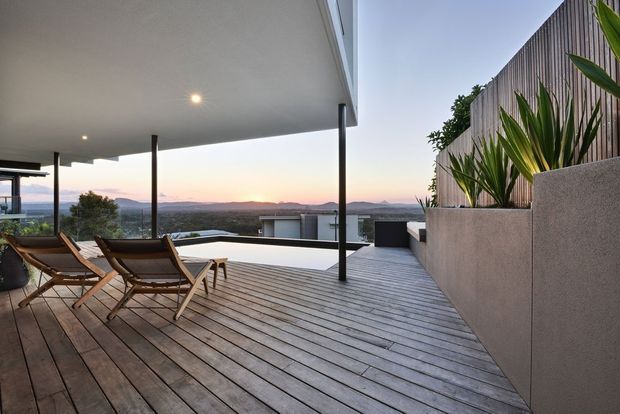 A modern rooftop or elevated outdoor terrace at dusk, featuring a broad timber deck, slim dark structural posts, a flat covered patio roof with recessed lighting, lounge chairs, and a raised concrete planter with tropical foliage. Beyond the terrace is a distant suburban or urban landscape with low hills.