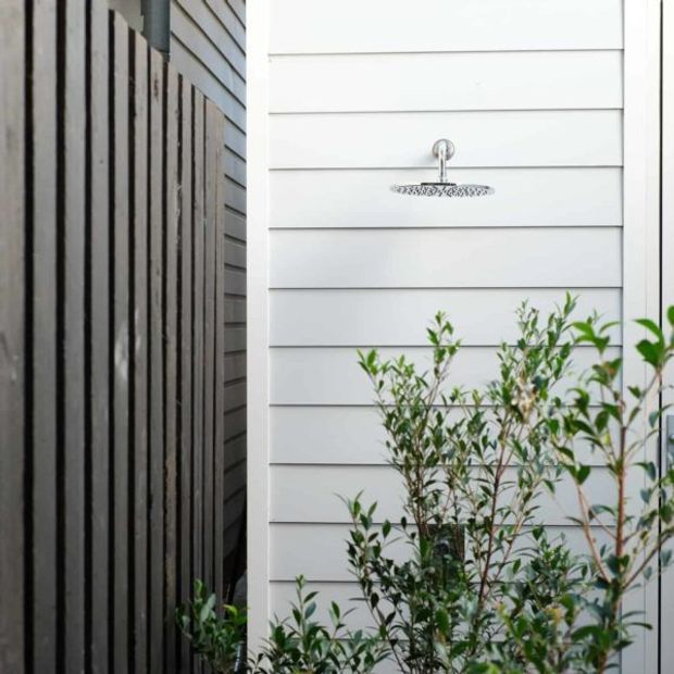 An exterior outdoor shower mounted on a white horizontal weatherboard-clad wall, with a round chrome showerhead and leafy planting in the foreground. A dark vertical timber privacy screen runs along the left side, creating a narrow landscaped shower area.