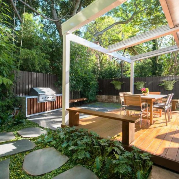 A bright landscaped backyard entertaining area with a timber deck, outdoor dining table and chairs, built-in barbecue, and a white-framed pergola with translucent or slatted roofing. Dense greenery, raised garden beds, stepping stones, dark timber fencing, and hanging planters create a secluded garden setting.