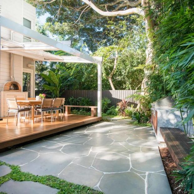 A bright, leafy outdoor entertaining area with a timber deck and dining setting beside a light-coloured home. The deck is sheltered by a slim white pergola, with a built-in outdoor pizza oven visible at the wall. Irregular natural stone paving extends through a lush landscaped courtyard, alongside a timber bench and dense planting.