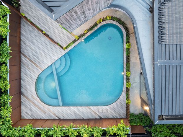 Aerial top-down view of a compact curved swimming pool surrounded by a timber-toned deck, stepped pool entry, planted borders, and adjacent grey roof and paved surfaces. The image focuses on the landscaped pool area and its integrated outdoor decking.