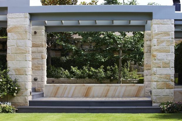 A landscaped outdoor courtyard or garden seating area framed by substantial pale stone pillars and a dark grey painted steel or timber overhead structure. The built-in bench is clad in light stone with natural veining, set against dense greenery, with dark tiled decking and manicured lawn in the foreground. The composition has clean contemporary lines combined with natural stone masonry.