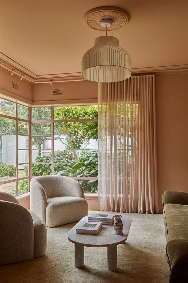 Warm, refined living room with large multi-pane corner windows overlooking a leafy garden. The room features peach-beige walls with ornate traditional ceiling cornices and a decorative ceiling medallion, a large pleated pendant light, full-height sheer curtains, curved upholstered armchairs and sofa, and a rounded stone coffee table on a pale carpet. The overall composition blends period architectural detailing with contemporary organic furniture and soft neutral furnishings.