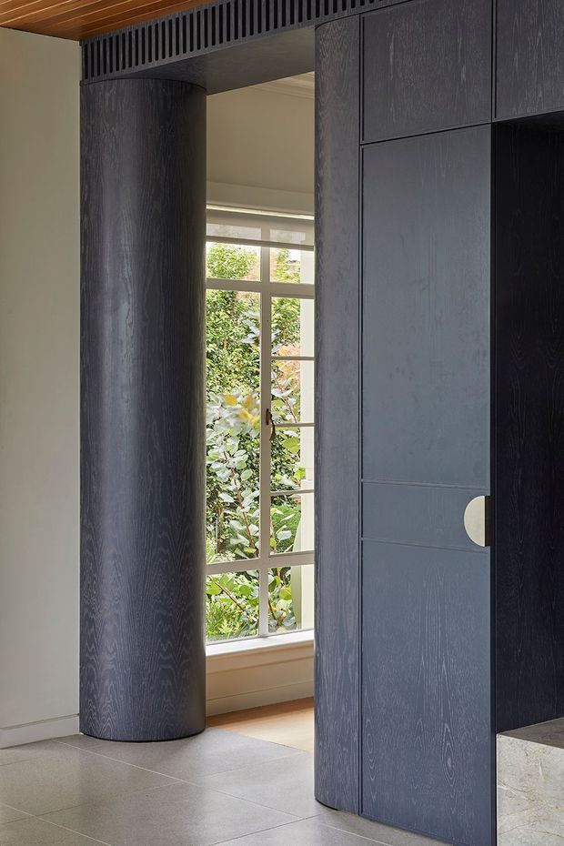 A refined interior passage featuring dark blue-grey timber veneer cabinetry and a matching curved cylindrical column, with a slatted timber ceiling detail above. A tall multi-pane window provides views to leafy planting outside, while large pale stone or porcelain floor tiles create contrast. The image emphasizes bespoke joinery, curved forms, and a concealed doorway with a round light-toned handle.
