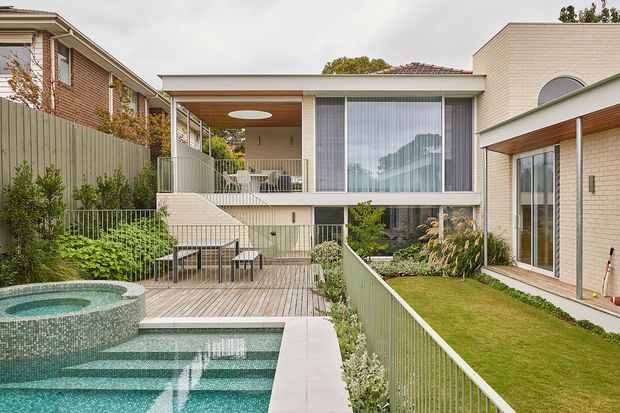 Exterior view of a contemporary multi-level residence arranged around a landscaped courtyard and pool. The composition shows pale brick walls, a low flat roofline, extensive floor-to-ceiling glazing, covered outdoor living areas, timber decking, planted garden beds, and a tiled swimming pool with a raised circular spa in the foreground.