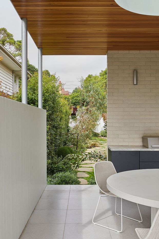 A covered outdoor dining and kitchen area opening onto a landscaped garden. The space features a warm timber-lined ceiling with a large circular skylight or roof opening, slim metal posts, pale tiled flooring, a light textured privacy screen, a pale masonry wall, dark built-in outdoor cabinetry with a barbecue, and a round white dining table with woven chairs. A stepping-stone garden path and dense planting are visible beyond.