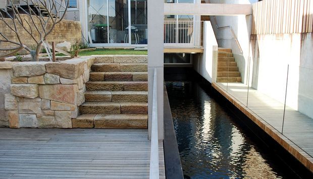A contemporary residential courtyard with a narrow linear reflecting pool or swimming pool running between white rendered walls and timber decking. Stone retaining walls and broad stone steps connect the lower deck to a glazed upper-level entrance, while a second stair rises beside the water. The composition combines clean modern architecture with rugged natural stone landscaping.