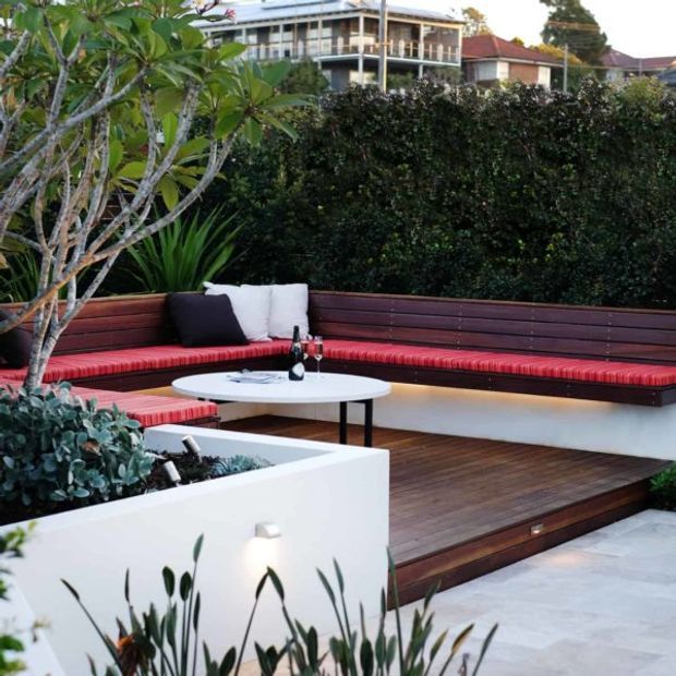 A landscaped outdoor entertaining area with a built-in L-shaped timber bench, vivid red cushions, a white round coffee table, timber decking, raised white planter boxes, and lush screening vegetation. Residential buildings are visible beyond the hedge.