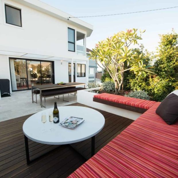 Contemporary outdoor entertaining terrace with dark timber decking, a large built-in L-shaped striped red lounge, round coffee table, dining table, and landscaped garden beside a white two-storey home with extensive glazing.