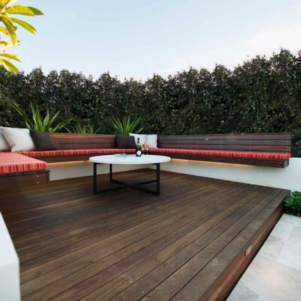 Outdoor terrace with a built-in L-shaped timber bench, red striped cushions, a small round coffee table, and dark timber decking. Tall hedging provides privacy, with tropical planting at the edges.