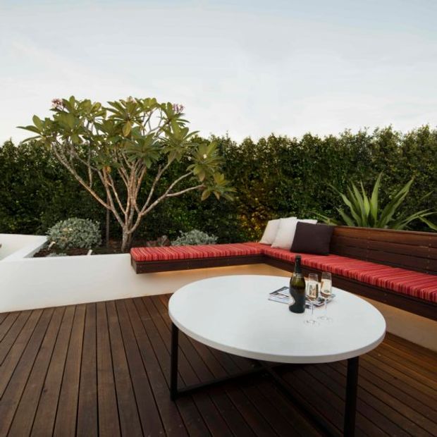 A landscaped outdoor terrace with dark timber decking, a built-in L-shaped bench with red upholstered cushions and dark timber backrest, white raised planters, and a round white outdoor table. Dense hedging and tropical-style planting create a private garden setting.