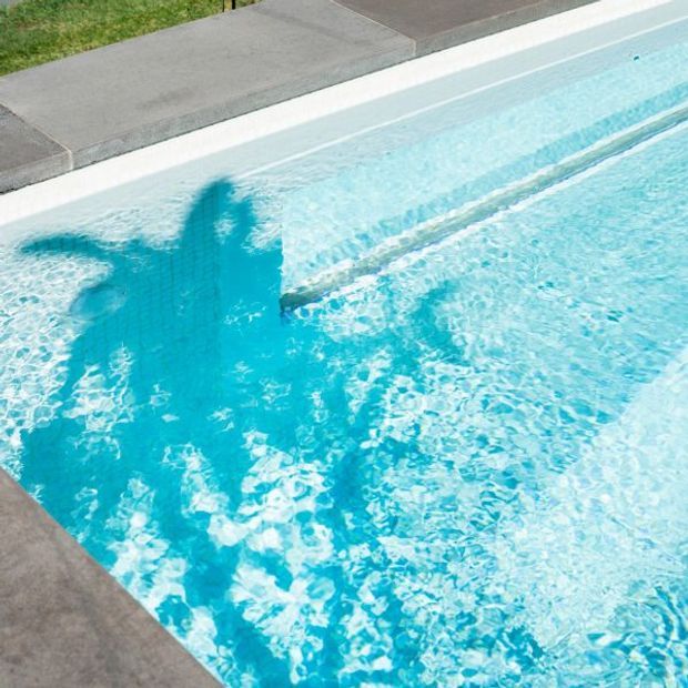 Close-up view of a bright turquoise swimming pool with gently rippling water, pale pool lining, and grey concrete coping around the edge. A narrow strip of lawn is visible beyond the pool.