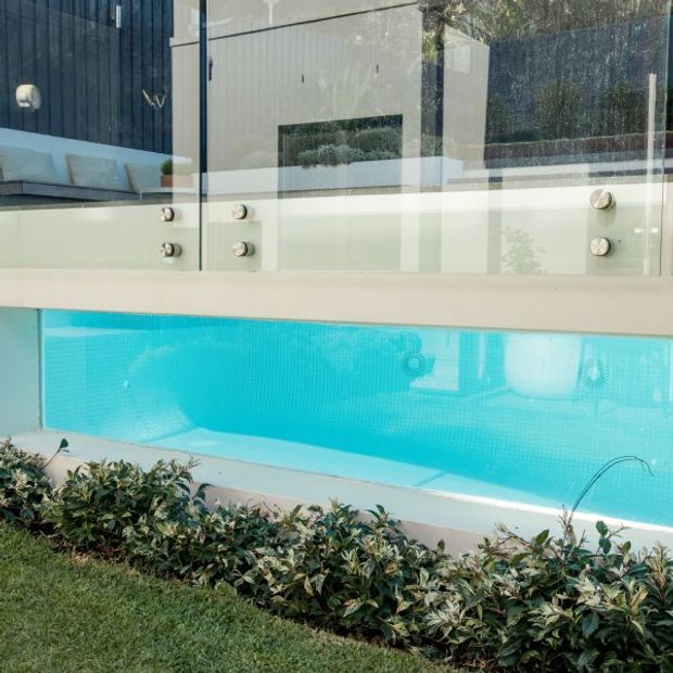 A bright turquoise swimming pool viewed from outside, with a white rendered pool surround and a clear glass balustrade secured by metal fittings. Reflections reveal a modern building and outdoor seating behind the glazing, while a planted garden bed and lawn border the pool in the foreground.