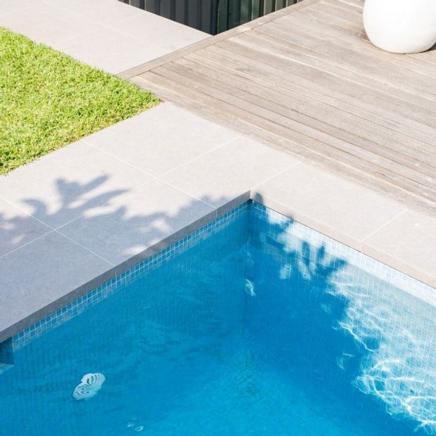 Close-up view of a bright outdoor swimming pool corner, with vivid blue tiled pool lining, pale large-format paving, and a weathered timber deck bordering a small lawn. Reflections and shadows from nearby planting fall across the water and paving.
