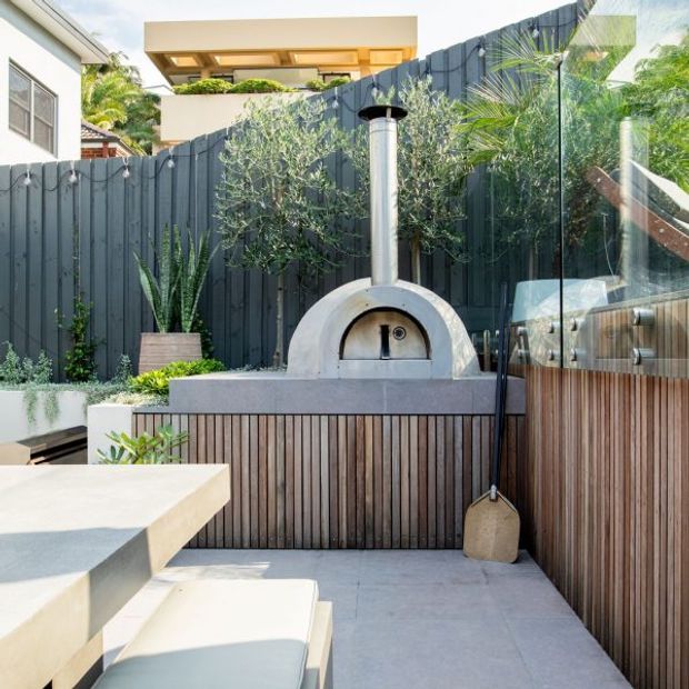 A bright contemporary outdoor entertaining area featuring a built-in wood-fired pizza oven with a cylindrical flue, set into a timber-slatted outdoor kitchen or cabinetry unit. Pale concrete paving and a matching dining table surround the oven, with a dark vertical privacy fence, glass screening, and landscaped potted planting. A modern residential building is visible beyond the fence.