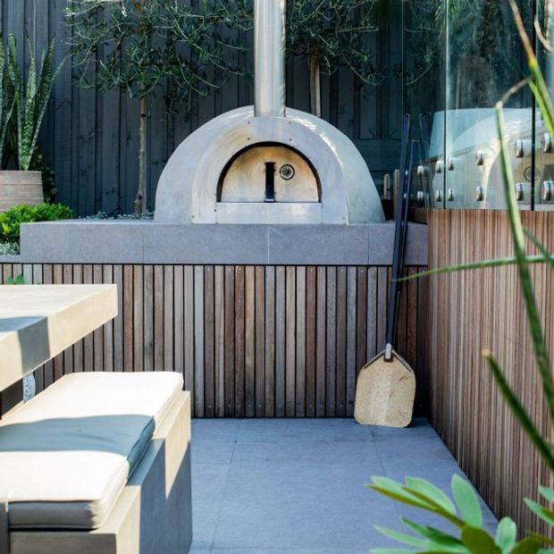 A compact outdoor entertaining area featuring a built-in dome pizza oven with a metal flue, set into a grey stone or concrete benchtop and surround. Vertical dark timber screening and timber-clad cabinetry frame the cooking zone, with large-format grey paving, white outdoor seating and surrounding planting. A stainless-steel outdoor appliance is visible to the right.