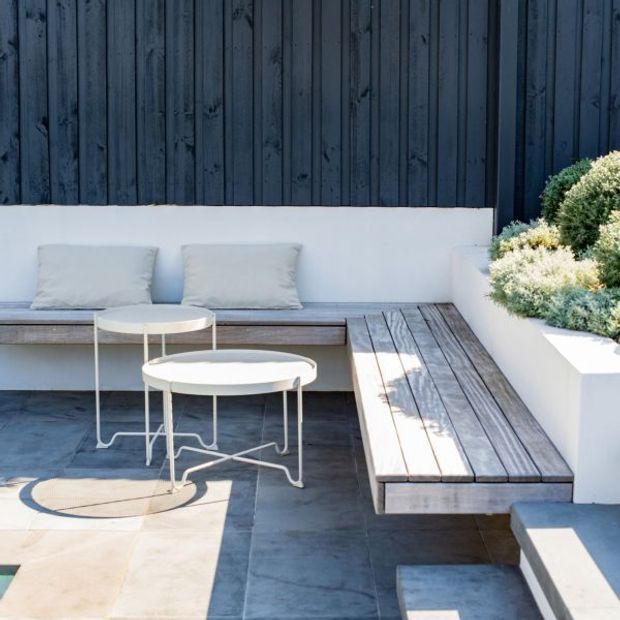 A compact outdoor courtyard or terrace seating area with built-in timber bench seating, pale cushions, two round white metal tables, dark tiled paving, and raised white planter beds. The space is framed by a dark vertically clad timber wall and low-maintenance greenery, creating a clean contemporary retreat.