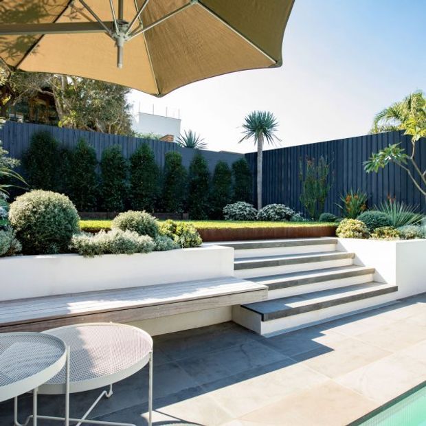A contemporary landscaped outdoor terrace with broad pale paving, stepped raised garden beds, built-in white bench seating, dark vertical privacy fencing, sculptural drought-tolerant planting, and a large tan cantilever-style umbrella. The edge of a swimming pool is visible in the foreground.