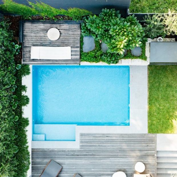 Aerial view of a landscaped residential pool area with a bright rectangular swimming pool, pale paved surrounds, extensive timber decking, built-in outdoor seating, lush planting, and a lawn edge. The composition emphasizes the pool and coordinated outdoor entertaining spaces.