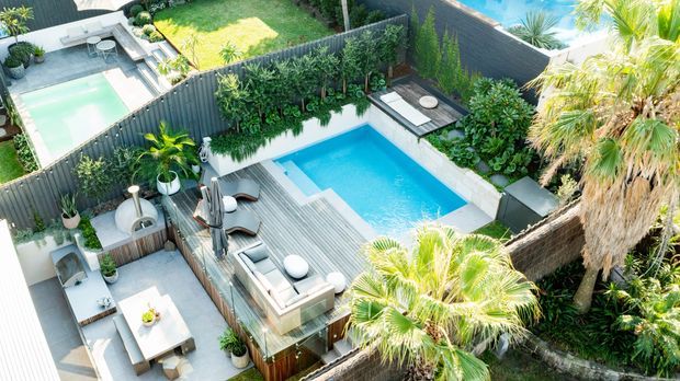 Aerial view of a landscaped residential outdoor entertaining area featuring a rectangular swimming pool, surrounding timber decking, outdoor lounge and dining furniture, privacy fencing, dense tropical planting, and adjacent lawns and paved areas.