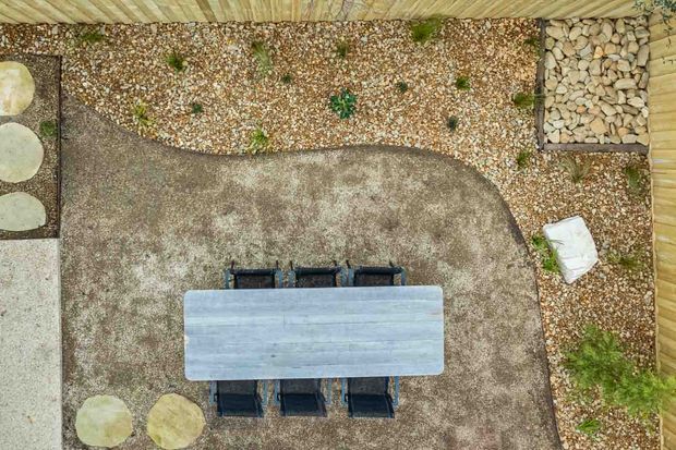 Overhead view of a landscaped outdoor dining area with a rectangular weathered timber dining table and dark chairs on a curved concrete or gravel-textured patio. Surrounding zones include pale pebbles, large stepping stones, planted pockets, and timber boundary fencing.