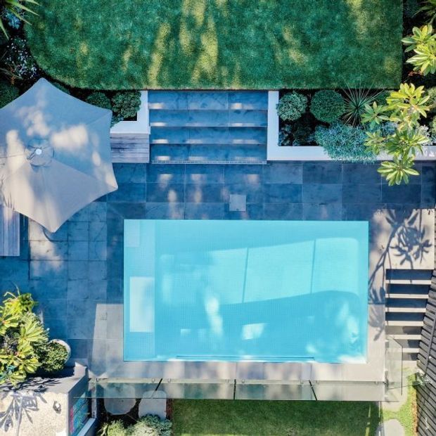 Aerial view of a landscaped residential pool area with a rectangular turquoise swimming pool, broad dark stone or concrete paving, a large outdoor umbrella, planted garden beds, steps, and a neatly clipped lawn. The composition is geometric and contemporary, with strong blue-green tones and dappled tree shadows.