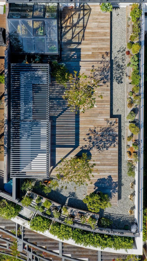 Aerial top-down view of a landscaped rooftop terrace with extensive timber decking, pale gravel garden beds, potted and planted greenery, and a large slatted pergola or screened outdoor structure. Strong linear shadows create a graphic pattern across the deck. The surrounding building edges and neighbouring urban structures are partially visible.