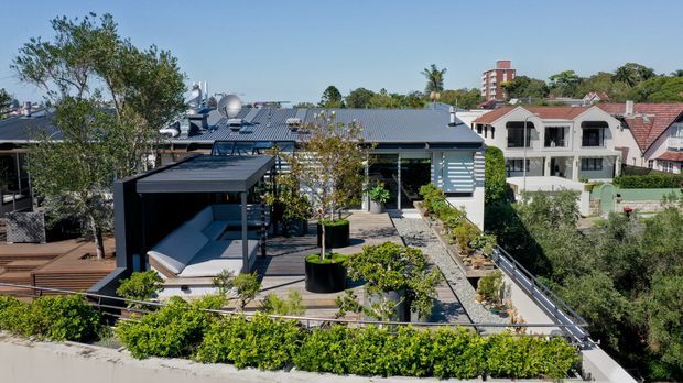 Elevated exterior view of a contemporary rooftop terrace and landscaped outdoor living area, with a dark-clad low-rise structure, covered seating pavilion, paved walkways, planter beds, and balcony railings. Surrounding residential buildings and mature greenery are visible in the background.