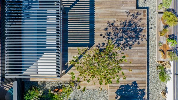Aerial view of a contemporary outdoor deck and landscaped courtyard, featuring broad timber decking, a dark slatted pergola or roof structure casting strong linear shadows, and gravel garden areas with planted greenery. The composition emphasizes geometric lines, natural timber, and contrasting dark metal elements.