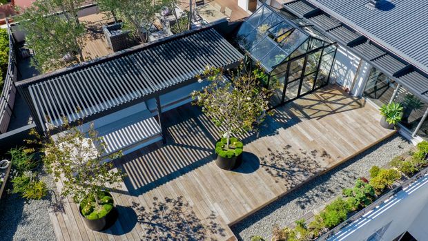 Aerial view of a contemporary rooftop terrace with extensive timber decking, dark corrugated roof structures, a glazed conservatory-like pavilion, outdoor furniture, potted trees, and landscaped gravel garden beds. Strong linear shadows and black-framed glazing create a modern, structured composition.