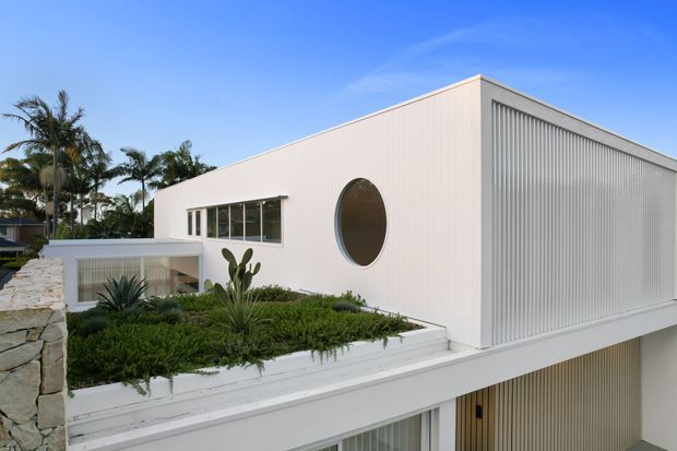 Exterior view of a clean-lined white modern building with a flat roof, vertical ribbed facade screening, a distinctive oval window, and extensive tropical landscaping on a rooftop or terrace level. A lower glazed and screened level is partially visible.