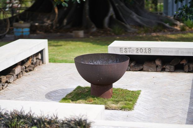 A landscaped outdoor courtyard or garden seating area with white concrete bench structures, stacked firewood stored beneath them, pale herringbone paving, and a central rusted metal fire bowl set into a small grass square. Mature trees and planting surround the space, creating a warm, textural outdoor gathering area.