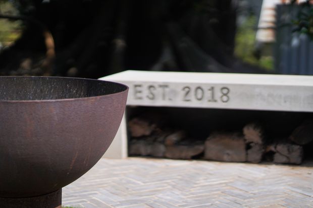 Close-up exterior view of a contemporary outdoor fireplace with a white rendered or concrete surround, stacked firewood in the opening, and a large rusted metal bowl fire pit in the foreground. The fireplace is set against a landscaped garden and paved with small rectangular tiles or bricks in a muted grey-beige palette.