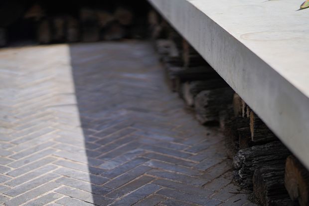 Close-up of an outdoor paved surface laid in a herringbone pattern beside a raised concrete slab or bench-like structure, with stacked natural timber or logs visible beneath it. The image is tightly framed and does not clearly establish the wider space.