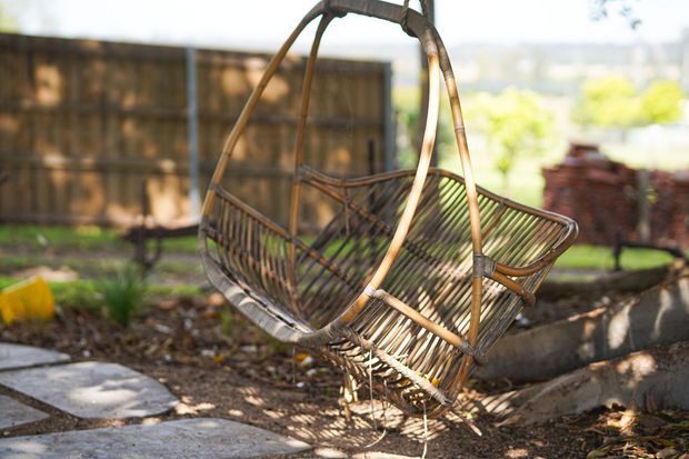 A rustic hanging egg chair made from woven natural wicker or rattan, suspended from a curved metal or timber frame in a garden setting. The chair sits beside a paved path and raised garden beds, with a timber privacy fence and greenery in the background. Warm natural light and earthy materials create a relaxed outdoor atmosphere.