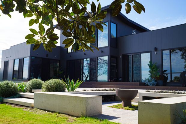 A contemporary dark-clad residential exterior with expansive floor-to-ceiling glazing, a projecting upper level, and a landscaped front courtyard. Concrete planter benches, clipped shrubs, ornamental grasses, and paved paths create a structured modern garden setting.