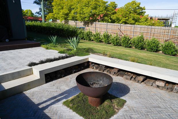 A landscaped outdoor courtyard or garden entertaining area with a circular metal fire bowl set into a paved patio. The patio uses light grey herringbone pavers and is edged by a built-in pale concrete bench or retaining wall with a dark stone-filled recess beneath. A lawn, clipped hedges, ornamental grasses and timber fencing form the background.