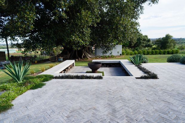 A landscaped outdoor courtyard or terrace with a broad pale grey herringbone-paved surface, a long rectangular reflecting pool or water feature, minimalist concrete seating, and a dark bowl-shaped fire or water feature. Mature trees, sculptural agave plants, clipped planting, and open rural landscape surround the space.