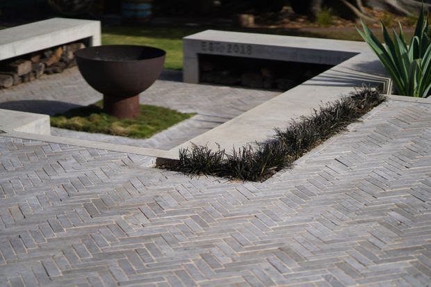 A landscaped outdoor courtyard or terrace paved in a light grey herringbone pattern, with raised concrete seating or retaining structures, integrated planting beds, a dark metal fire bowl, and large architectural succulents. The design has a minimalist, geometric character.