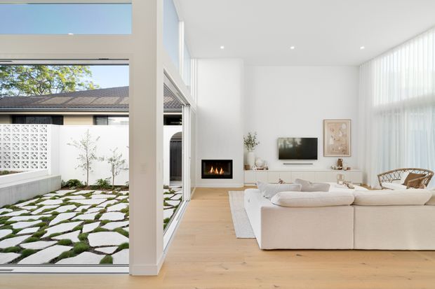 Bright contemporary living room with a large white sectional sofa, pale timber flooring, white walls, a linear fireplace, minimalist media console, and floor-to-ceiling glazing. The glazing opens to a landscaped courtyard with stepping stones, moss, a breeze-block privacy screen, and a water feature or narrow pool edge. A high-level horizontal window and full-height sheer blinds enhance the airy interior.