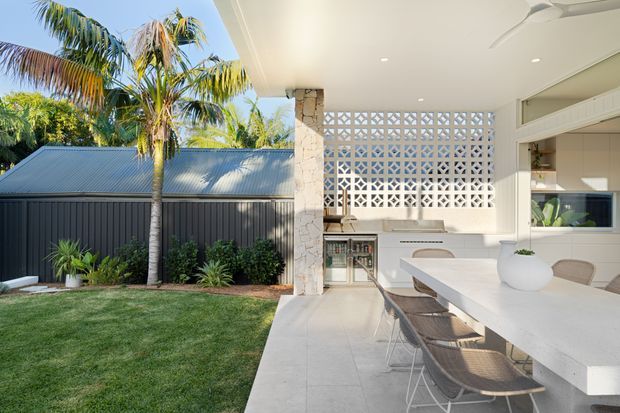 A bright contemporary outdoor kitchen and dining area opening onto a landscaped backyard. The space features a large pale stone island with integrated seating, a white tiled or rendered floor, white cabinetry, a decorative breeze-block screen, a stone-clad column, and a corrugated metal roof visible over the adjacent structure. Palm trees and lawn create a tropical garden setting, while a ceiling fan and recessed lighting support outdoor entertaining.