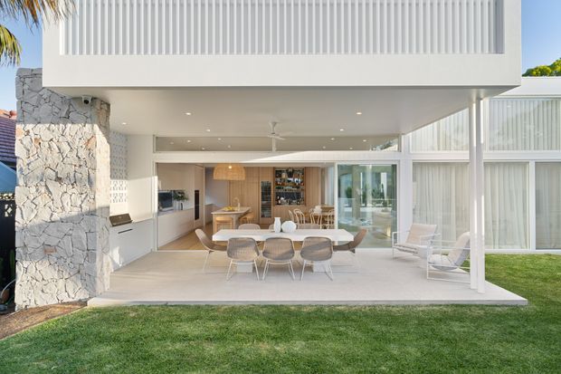 Bright contemporary two-storey residential extension viewed from the garden, with a sheltered outdoor dining terrace flowing into an open-plan kitchen and dining area. The facade combines white vertical screening and extensive glazing with a rough natural-stone structural wall. Inside, light timber cabinetry, a dining table, bar joinery and neutral furnishings are visible, while the terrace includes lounge seating and a white ceiling fan.