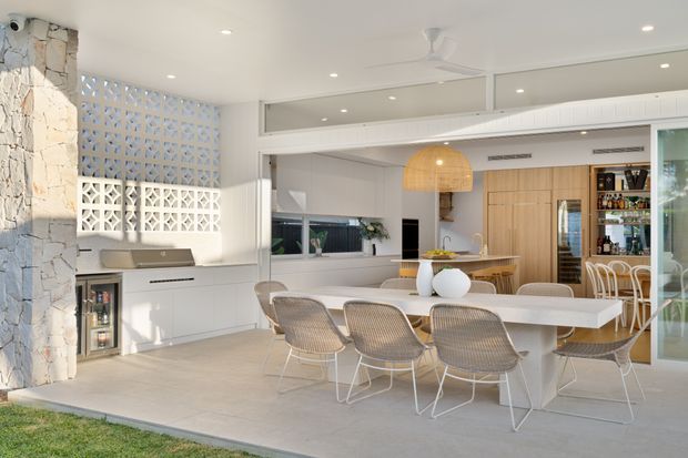 Bright open-plan coastal outdoor entertaining area with a dining table, woven dining chairs, white built-in cabinetry, a compact kitchen/bar area, stone feature cladding, breeze-block screens, and a covered ceiling with recessed lighting and fan. The space opens directly to a lawn through large glazed doors.