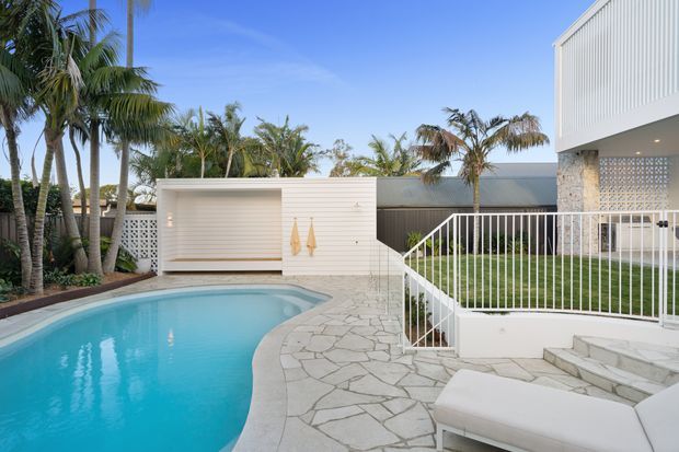 A bright tropical residential pool area beside a modern coastal-style house. The image shows a curved swimming pool, irregular natural-stone paving, white metal balustrades and steps, a white poolside storage or outdoor entertaining structure, patterned breeze-block screening, and abundant palm landscaping.
