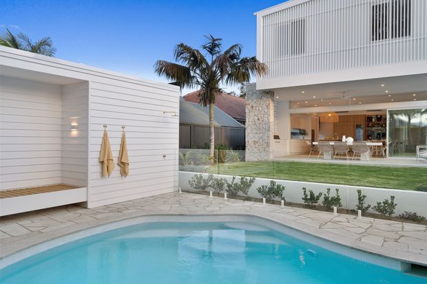 A bright contemporary coastal home viewed from a swimming pool courtyard. The scene features a turquoise pool, pale stone paving, a white horizontal-clad poolside structure with an outdoor shower or towel area, landscaped lawn and planting, and a two-storey white house with vertical screening, a natural stone feature wall, and expansive glazed living areas opening to the courtyard.