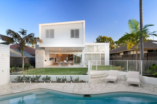 Exterior view of a contemporary white multi-level residence with a strong rectangular form, vertical screening, open-plan glazing, a landscaped lawn, paved pool terrace, lounge chairs, and a swimming pool in the foreground. Palm trees and neighboring suburban homes frame the property.
