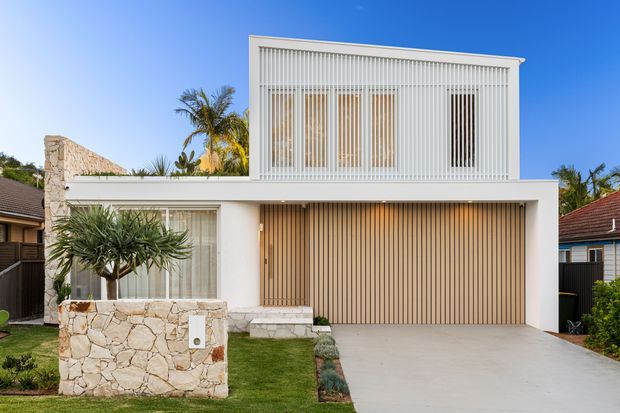 Front exterior view of a contemporary two-storey residence with a crisp white rendered facade, upper-level vertical screening, warm timber-toned vertical garage and entry cladding, large glazed openings, and a pale natural-stone front planter wall. A broad concrete driveway and landscaped front garden complete the street-facing composition.